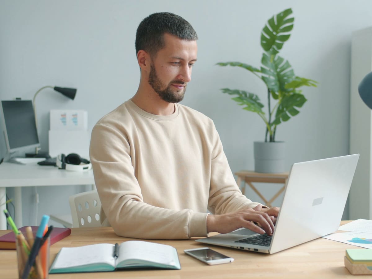 A man in a cream sweater types at a silver laptop at a wooden desk in a light home office, an open notebook, pens and papers beside him and a tall plant behind.